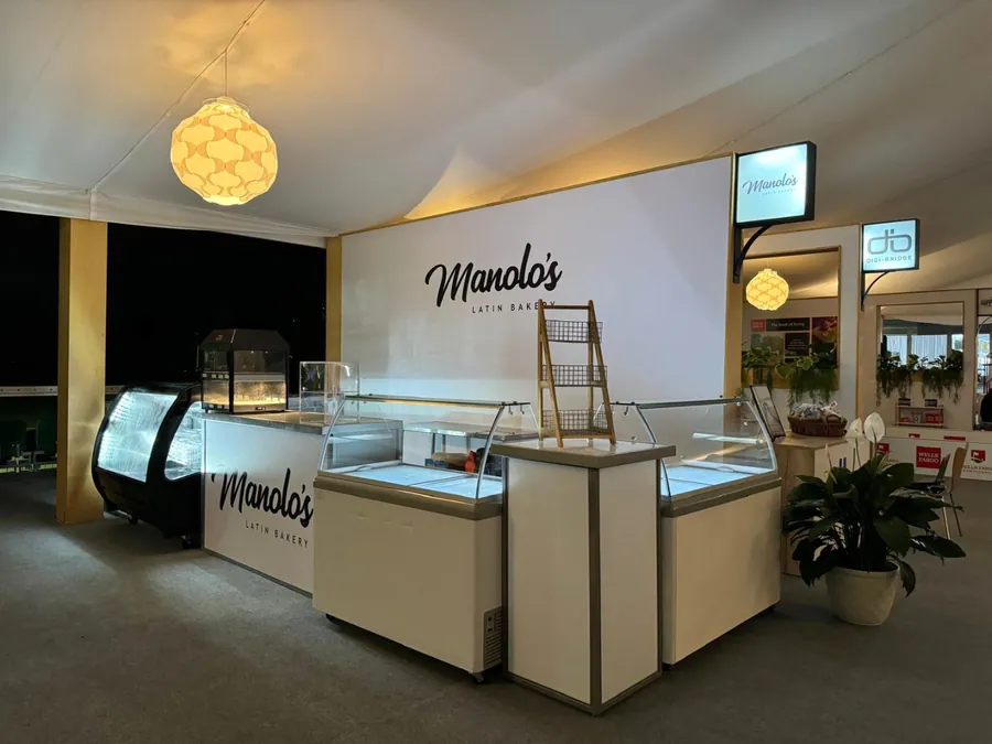 The completed bakery environment at night, with illuminated glass display cases, a warm-lit back bar, branded fascia, and planting at the counter ends.