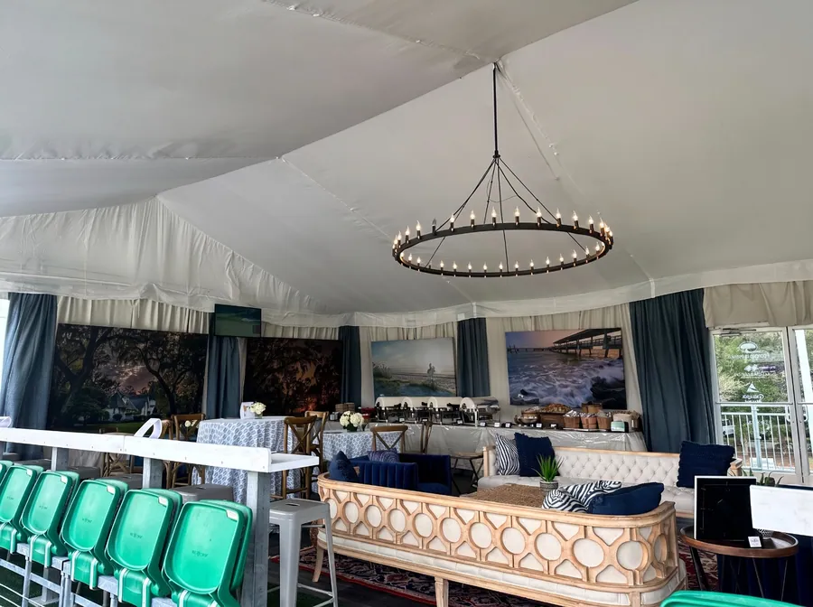 A tented hospitality interior with a chandelier, patterned banquettes, framed landscape prints, and a raised seating platform above stadium seats.