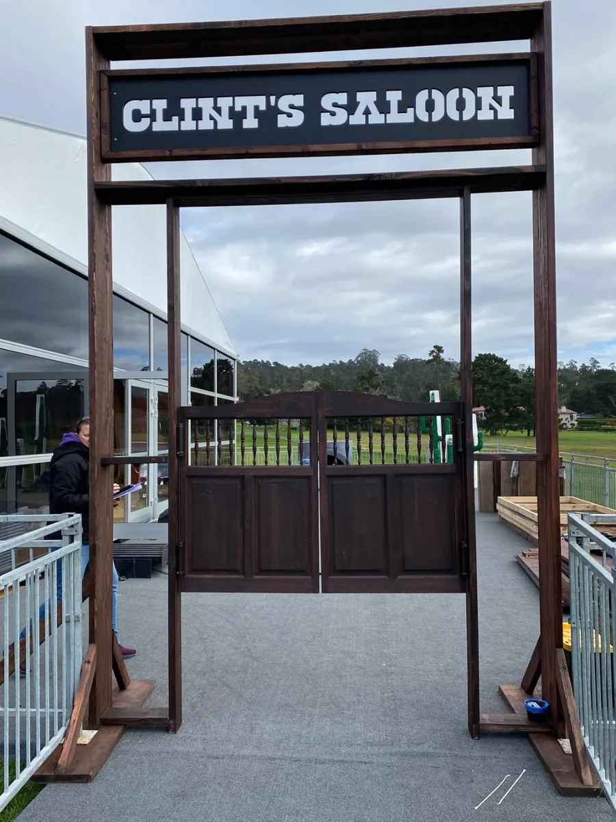 A dark timber saloon-style entry frame with a lit sign band and swing gates, opening onto a course beyond.