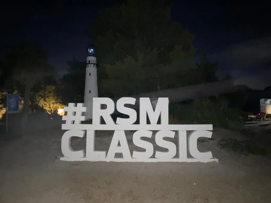 Large three-dimensional letters spelling a tournament hashtag, lit at night in front of a lighthouse.