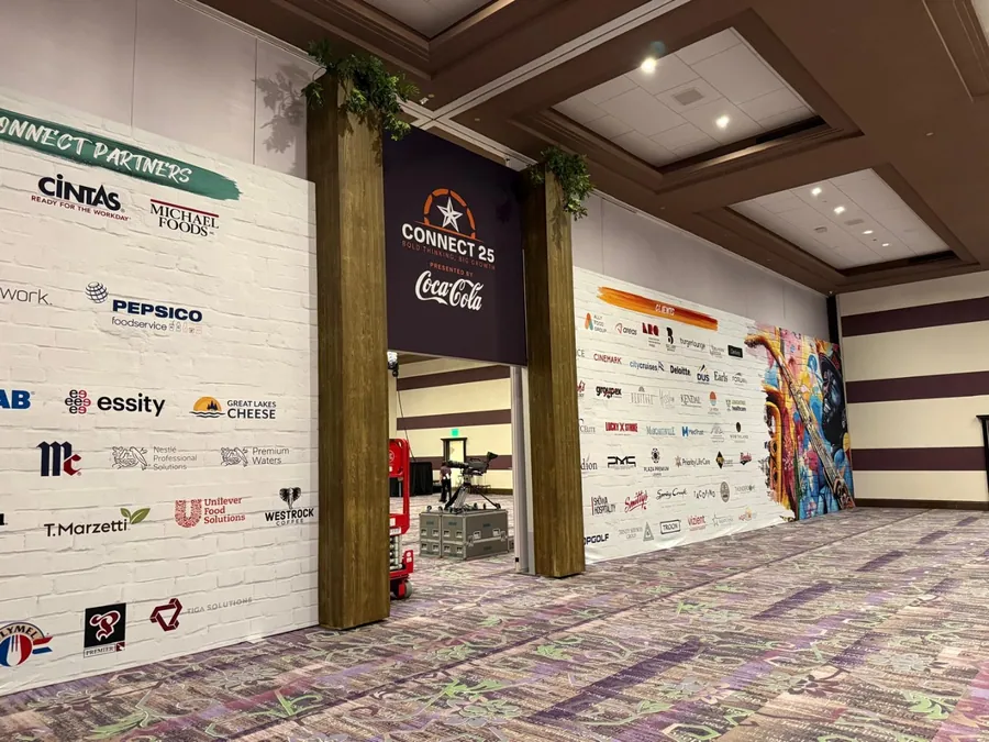 A long interior corridor lined with a white sponsor wall carrying dozens of partner marks, framed by timber portals and greenery.