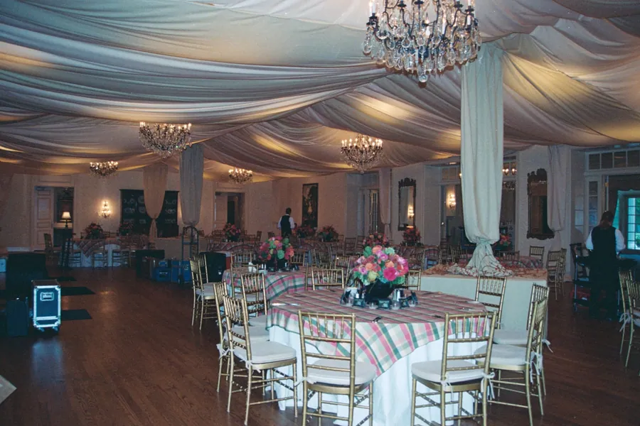 A ballroom draped in swagged white fabric with chandeliers, gold chairs, and pink floral centrepieces on round tables.