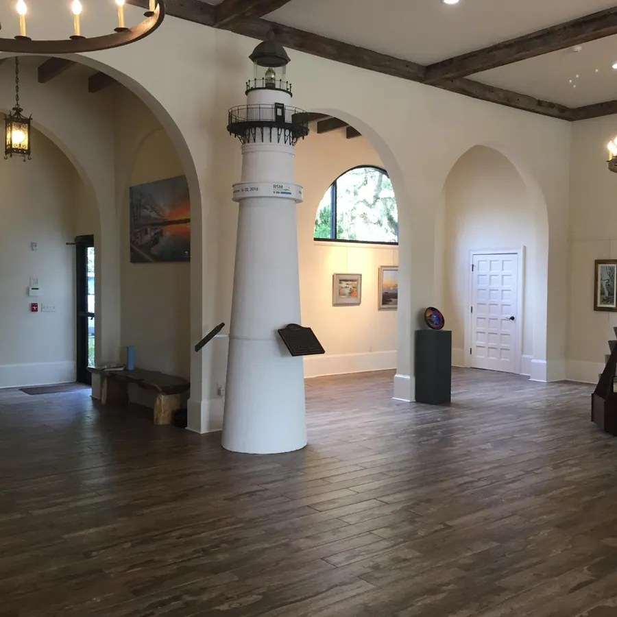 A tall white interior with a full-height lighthouse replica, arched windows, timber floors, and framed artwork on the walls.
