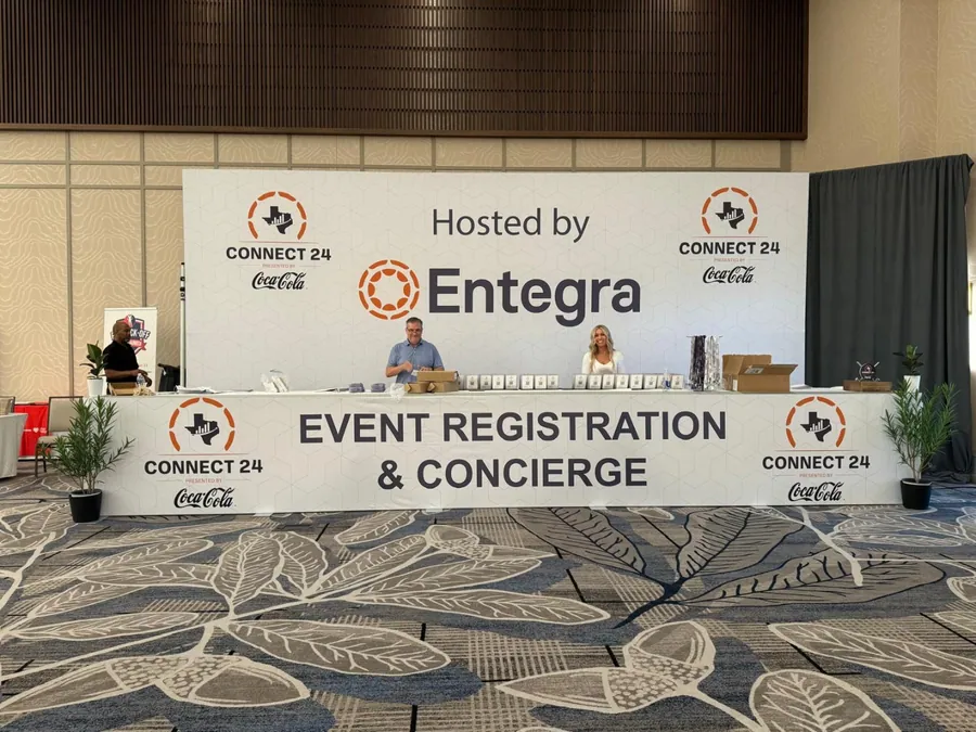 A branded registration and concierge desk with a printed header wall and repeat partner marks, staffed inside a function room.